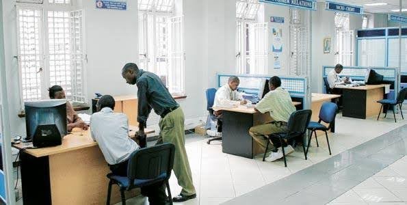 People interacting with office staff at service desks inside a bright bank or customer service hall.